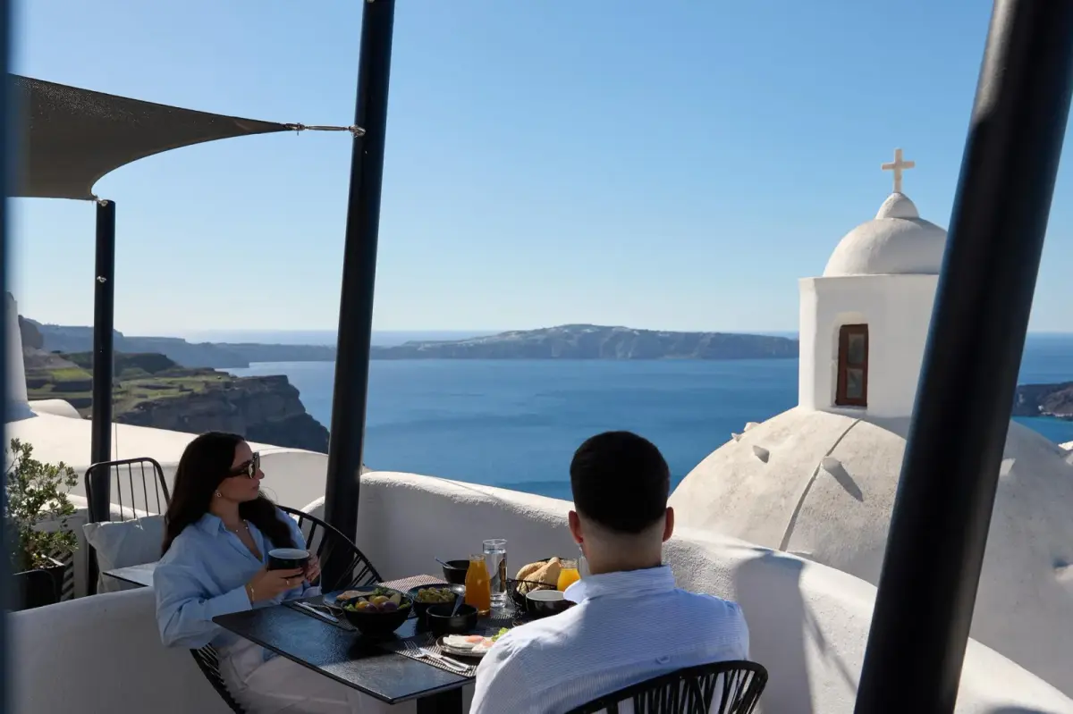 A couple at breakfast on a terrace at Aroma Suites, a church dome and the caldera behind