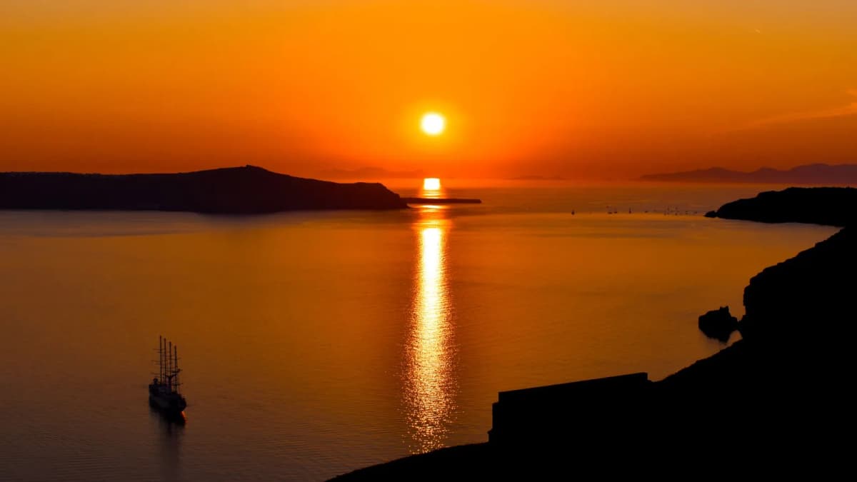Oia sunset over the Santorini caldera as the sun drops behind the volcanic islands with a sailboat in the bay