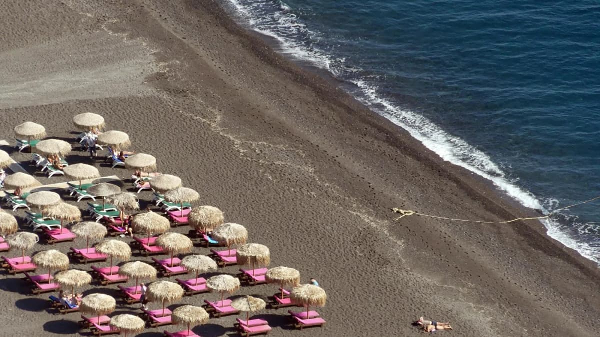 Sunbeds and straw umbrellas on black volcanic sand beach in Santorini with Aegean Sea waves