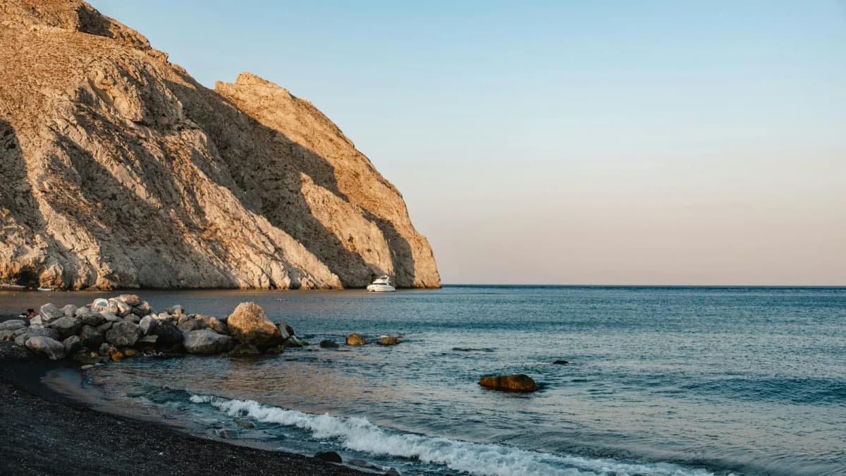 Volcanic white cliffs rising above dark sand beach at Vlychada, Santorini with blue Aegean water