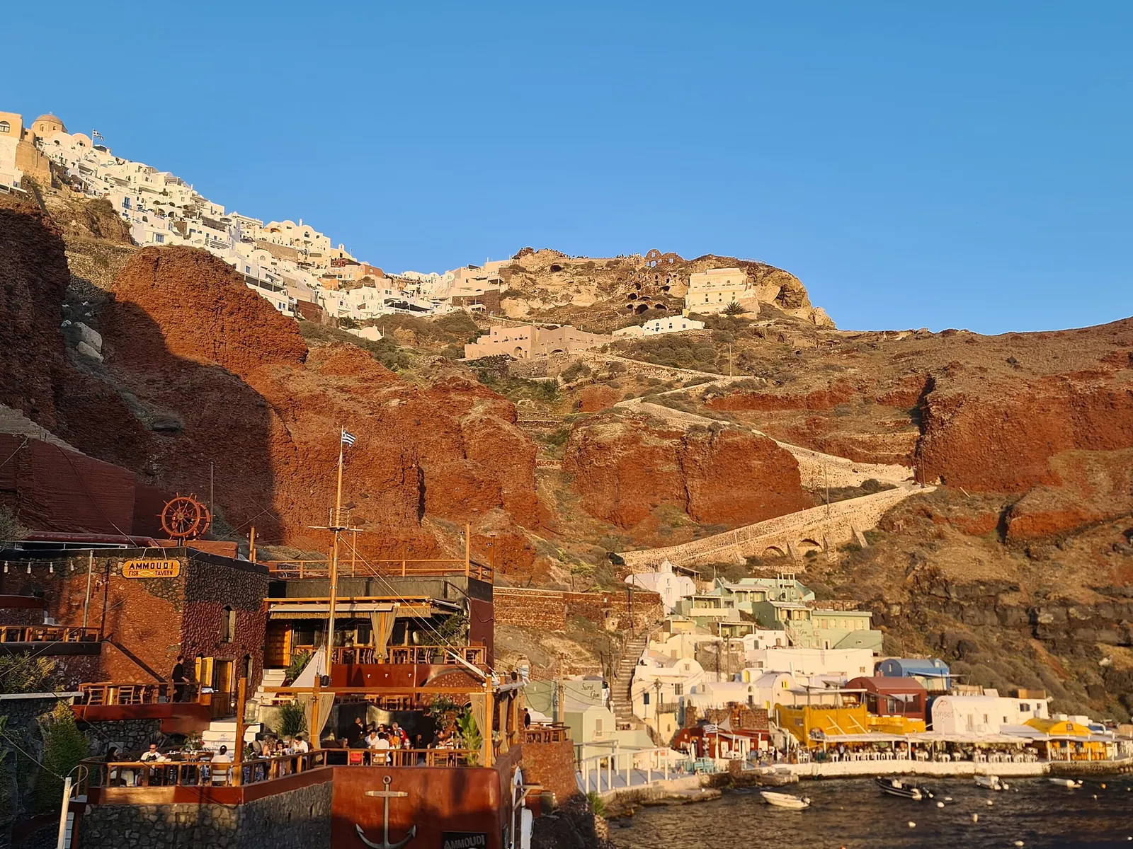 Waterfront tavernas at Ammoudi harbour in evening light, Oia on the cliff above