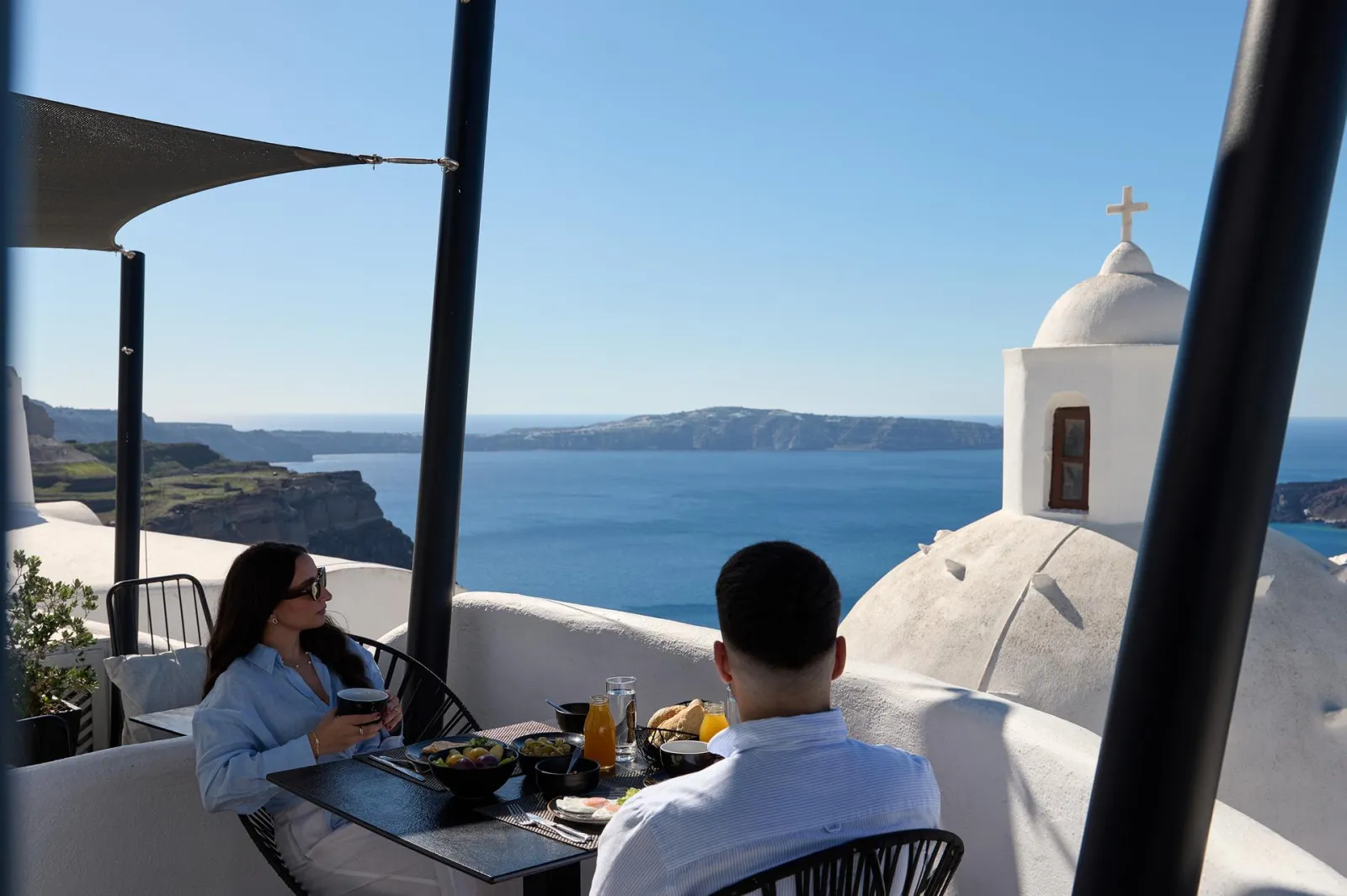 A couple at breakfast on a terrace at Aroma Suites, a church dome and the caldera behind