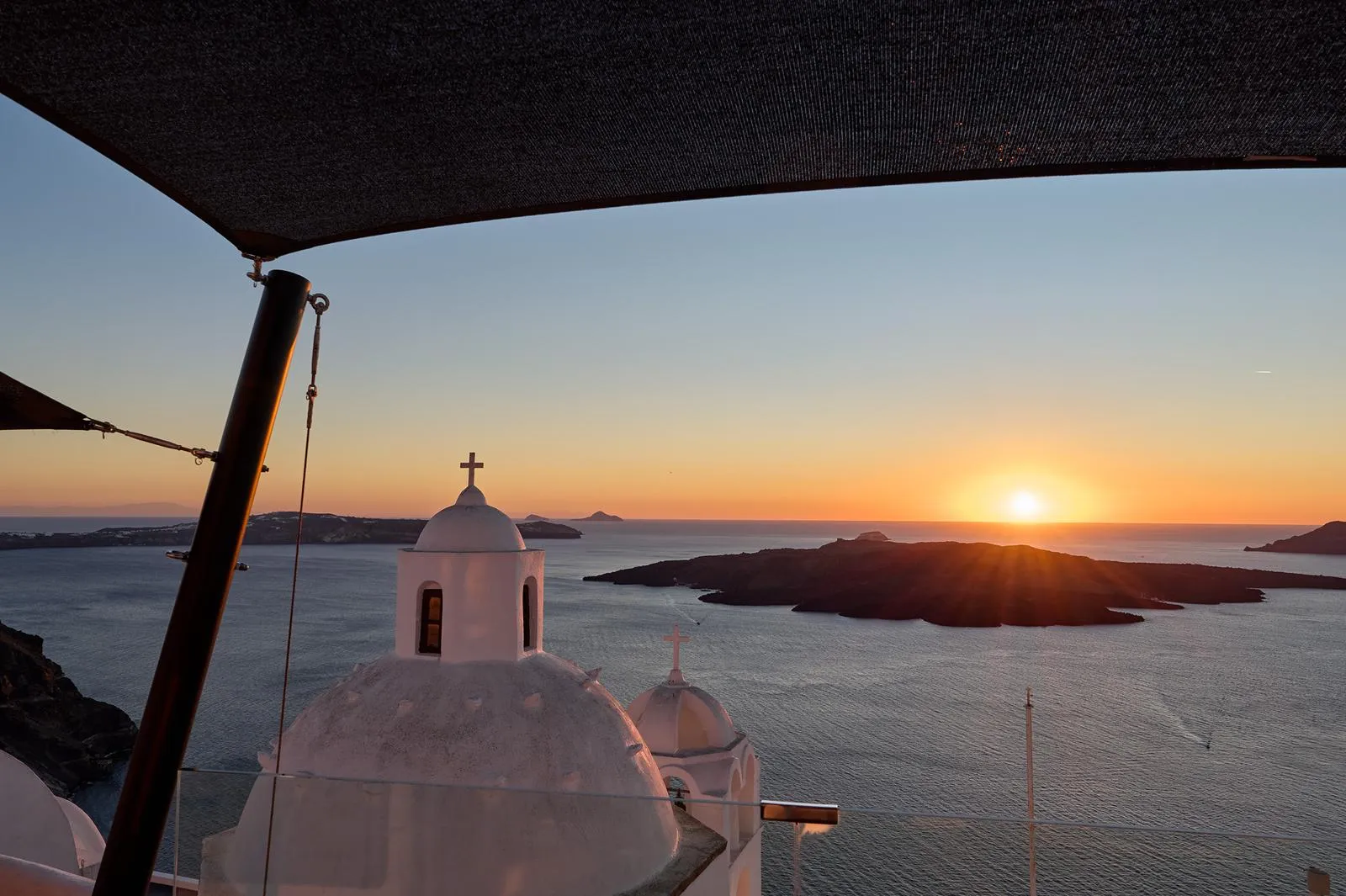 Sunset over the caldera and Nea Kameni from a shaded terrace at Aroma Suites, church domes below