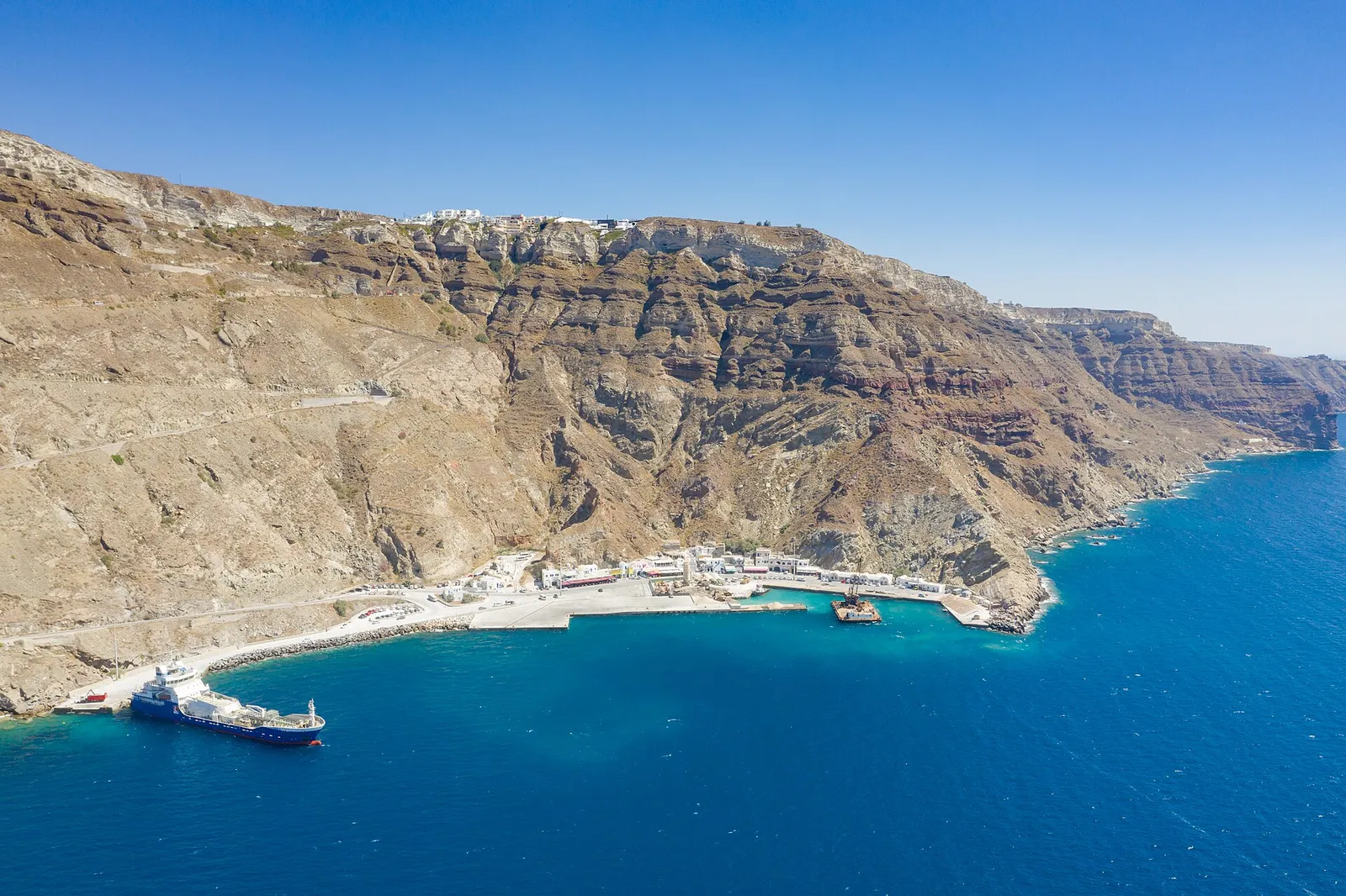 Aerial view of Athinios port at the foot of Santorini's cliffs, with the hairpin road climbing towards the caldera rim
