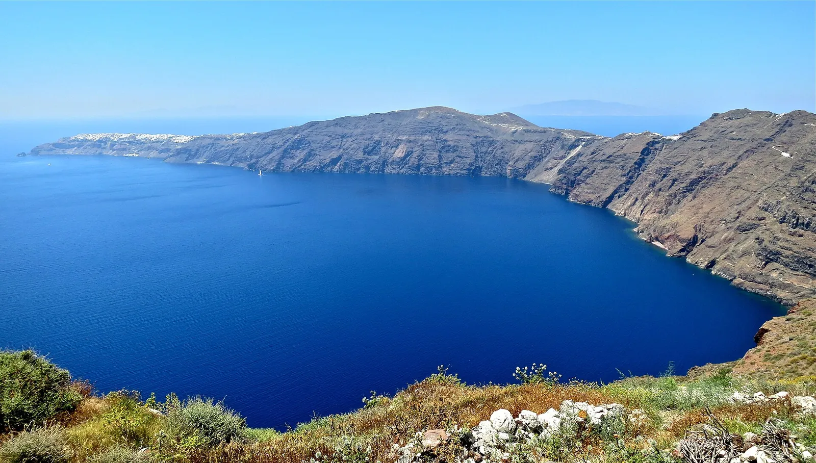 The caldera rim and deep blue water seen from the hiking trail, green scrub in the foreground