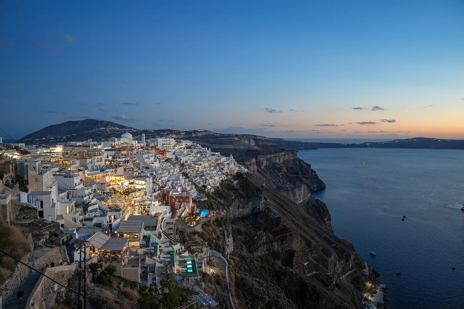 Fira's lights coming on along the caldera edge at dusk, the cliff dropping to the sea