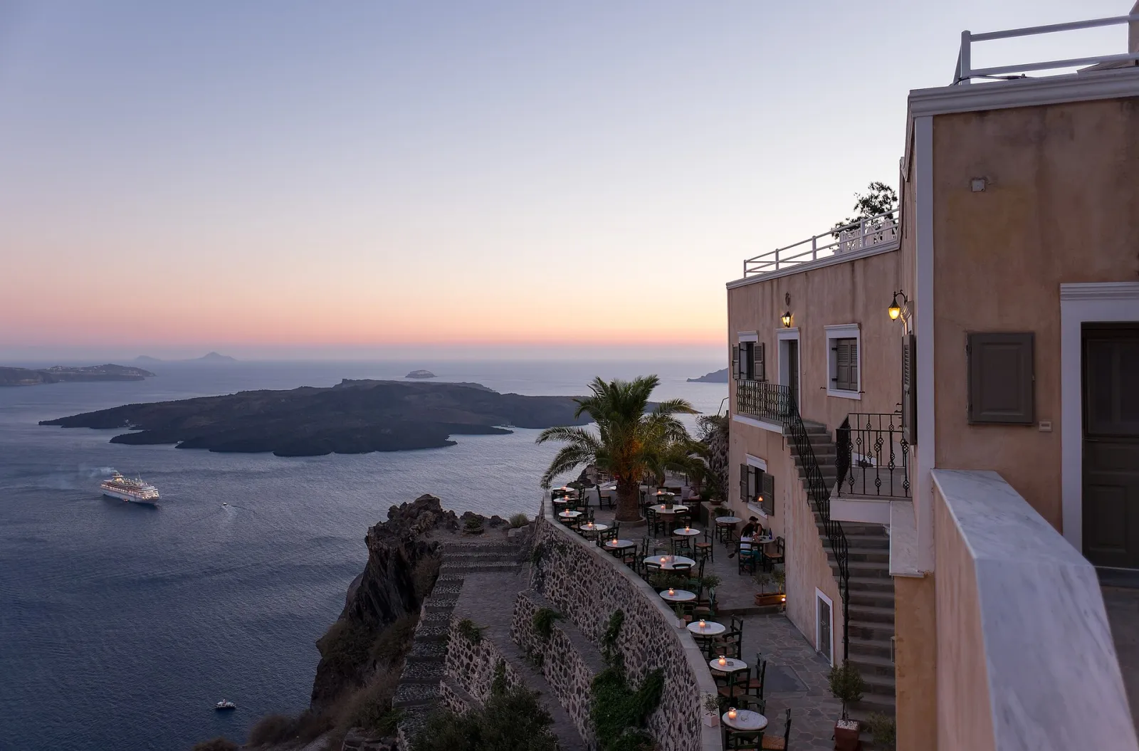 Restaurant terrace on the caldera edge in Fira at dusk, with Nea Kameni and a cruise ship beyond