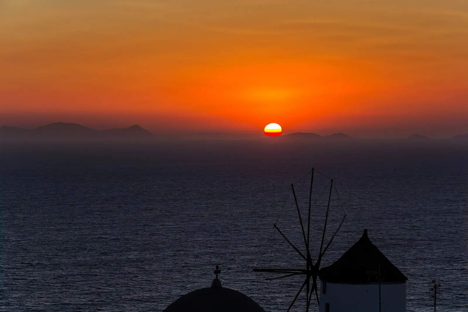 Sun setting over the sea at Oia with a windmill and church dome in silhouette