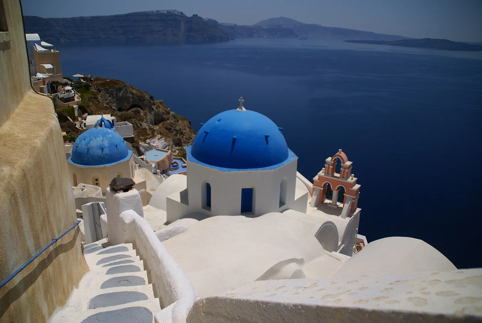 Two blue domes and a bell tower in Oia, with whitewashed steps leading down towards the caldera