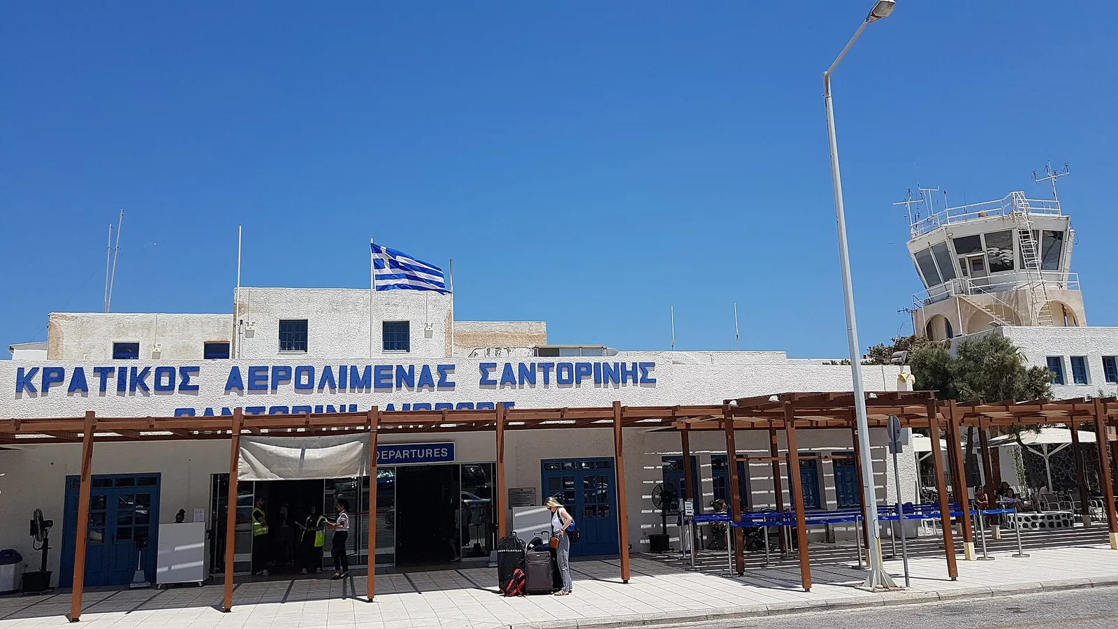 Entrance of the Santorini airport terminal under a Greek flag, with the control tower behind