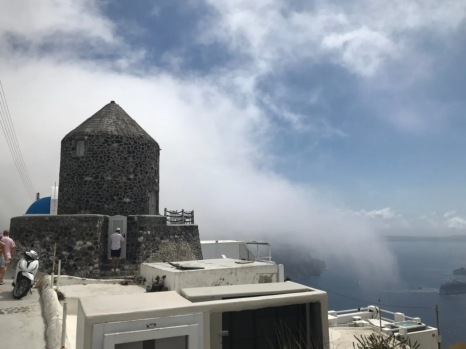 Old stone windmill beside the caldera path before Imerovigli, cloud rolling over the cliff