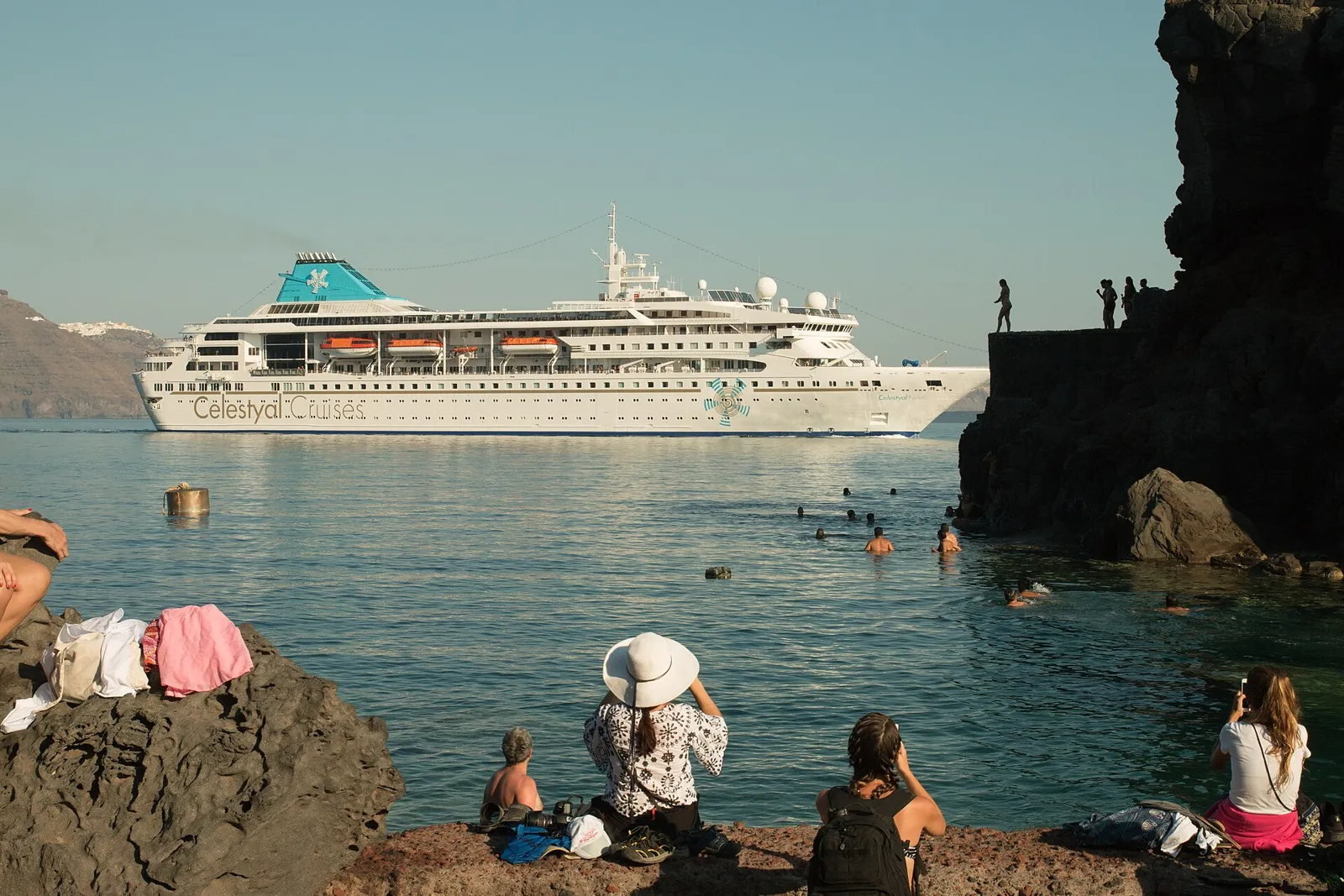 People swimming off the rocks at Agios Nikolaos islet near Ammoudi, a cruise ship passing